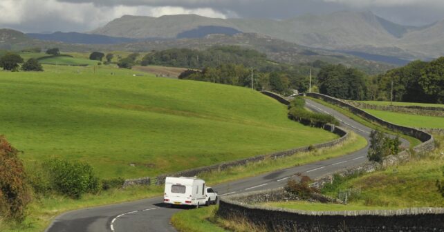 Car towing a caravan on a winding road through green hills in England.